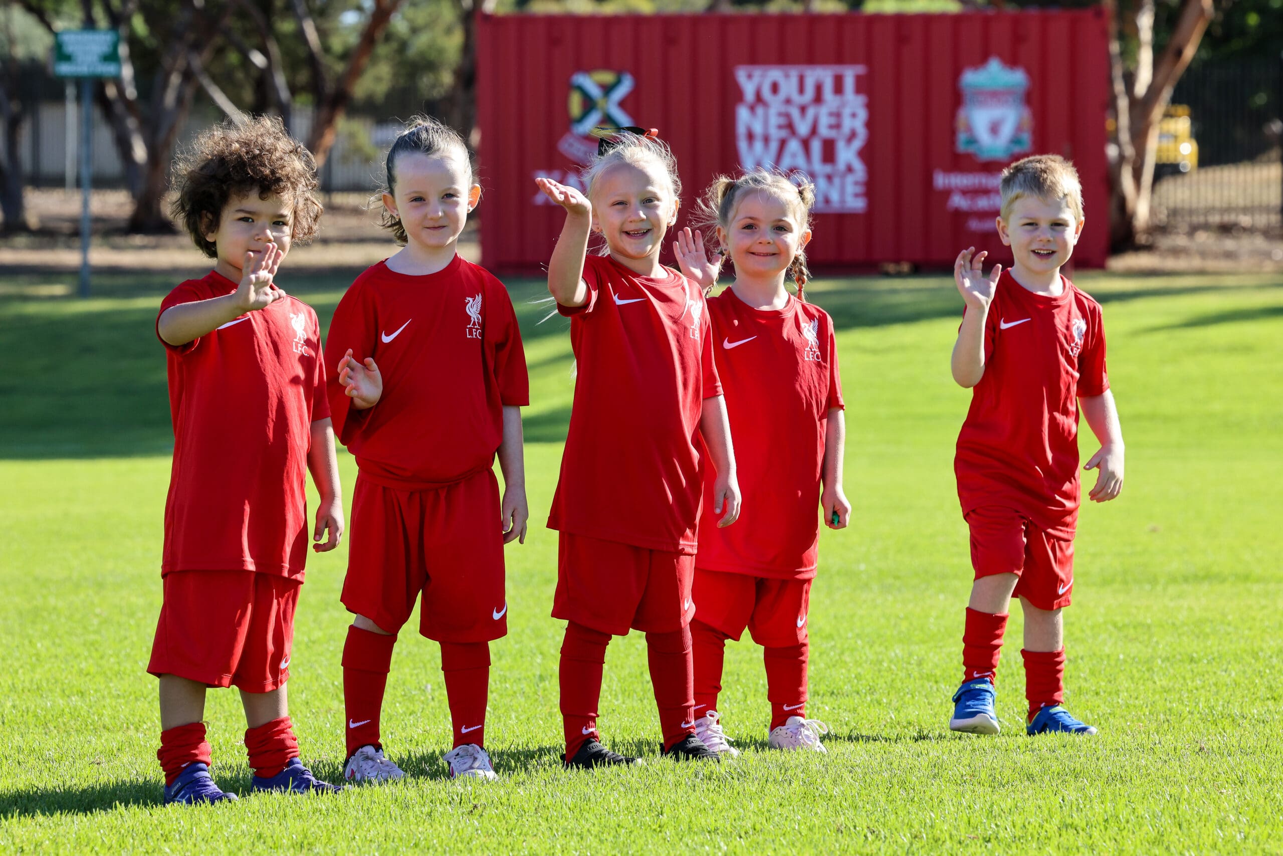 Early years students wearing Liverpool uniforms waving at the camera.