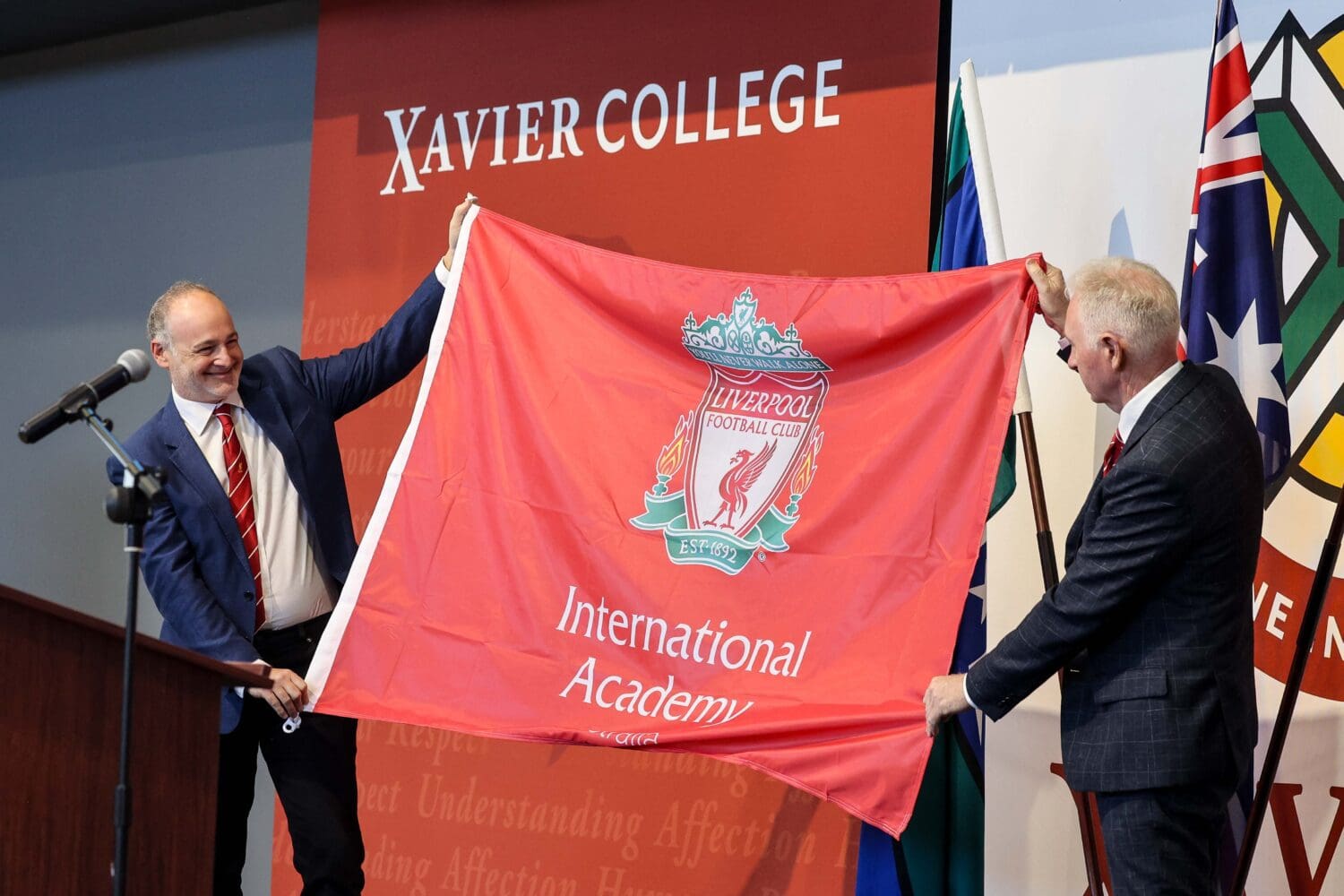 Lo-Res.LFC&XAVIERCOLLEGEPARTNERSHIP-113 Kevin Kalinko, Director of LFC International Academy, and Mark Flaherty, Principal of Xavier College, standing on stage holding a Liverpool International Academy flag between them.